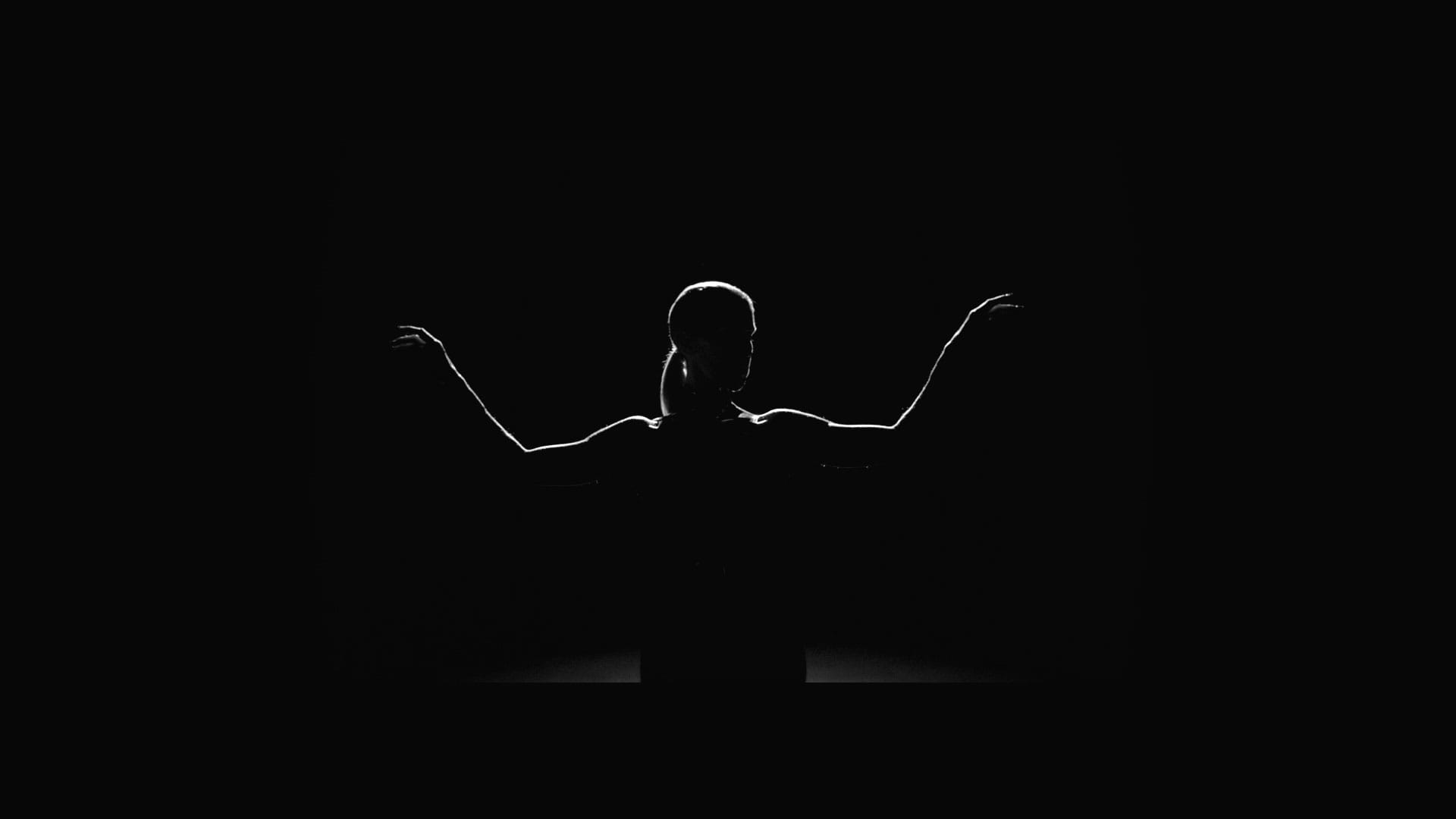 Viewed from behind, a single spotlight traces the turned head and outstretched arms of a woman on a darkened stage.